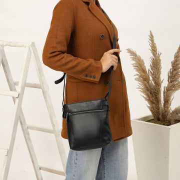 Person wearing a brown coat holding a black crossbody bag in a minimalistic setting with a plant.
