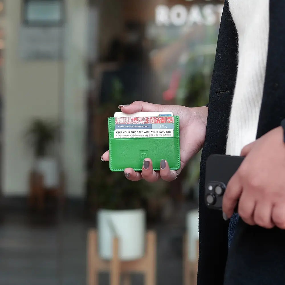 A person holding a green leather card holder wallet with a card partially inserted, photographed in a real indoor environment with a blurred background.