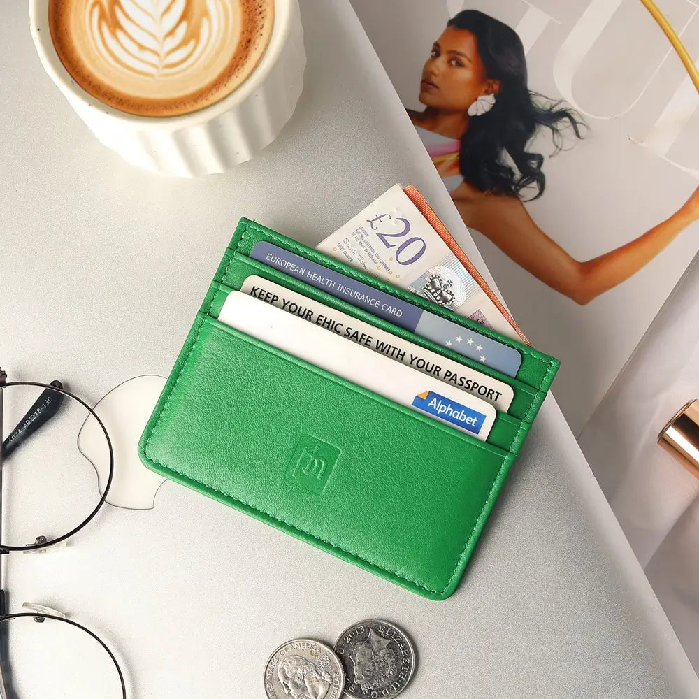 A green leather card holder wallet with multiple card slots, shown open with cards and cash partially visible, styled on a tabletop with a coffee cup, eyeglasses, coins, and a magazine.