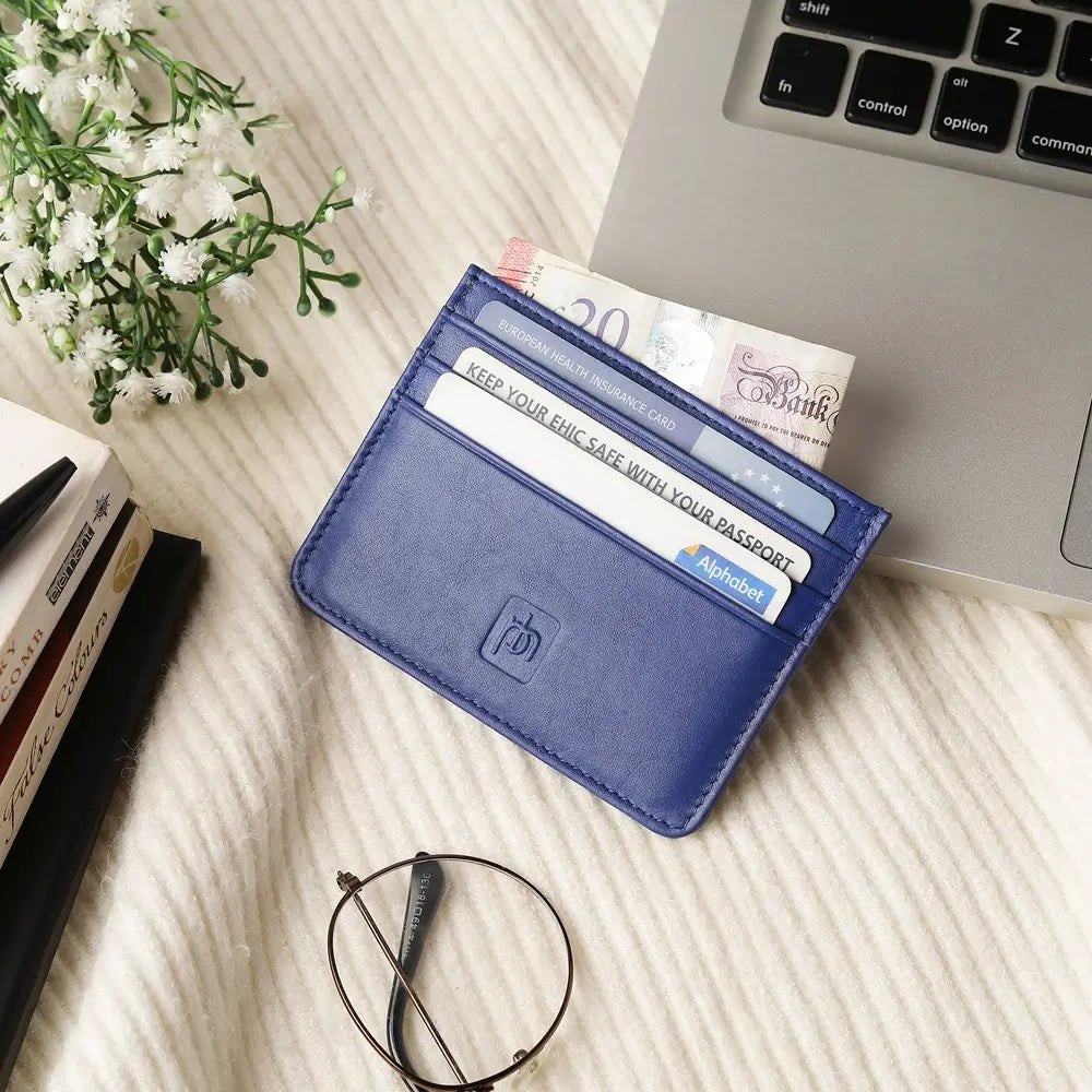 A navy slim card holder wallet with multiple card slots, shown holding credit cards and cash on a light textured fabric surface next to a laptop, books, glasses, and small white flowers.