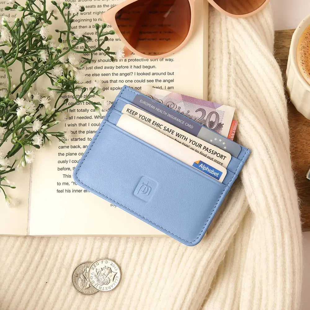 A pastel blue leather card holder wallet with multiple cards inserted, styled on a cream knit blanket over an open book with flowers, cups, and a coin.
