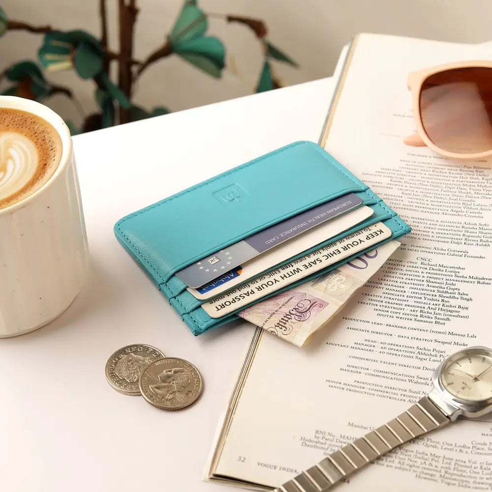 A turquoise blue leather card holder wallet with multiple cards and cash, styled on a white tabletop with a coffee cup, sunglasses, coins, an open book, and a watch.