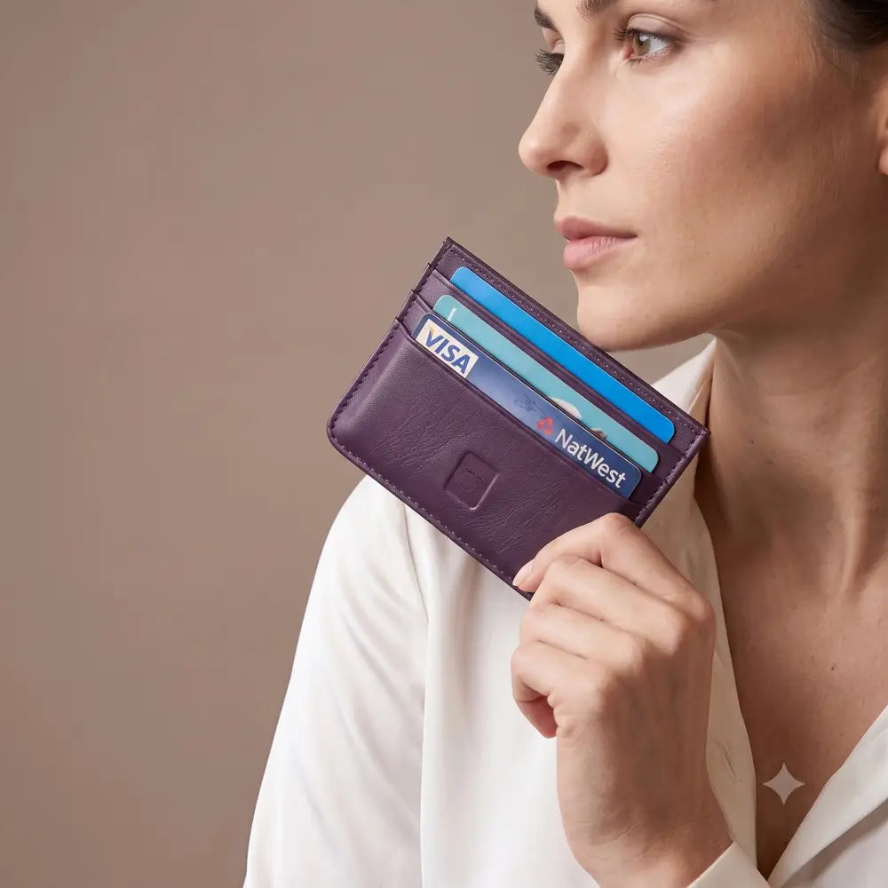 A woman in a white blouse holds a slim purple leather cardholder with several credit cards inserted, photographed against a plain taupe background.