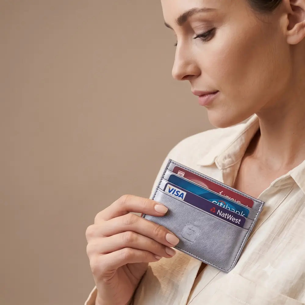 A woman in a cream-colored blouse holds a slim sliver leather cardholder wallet containing several credit cards, including Visa and NatWest cards, against a solid beige background.