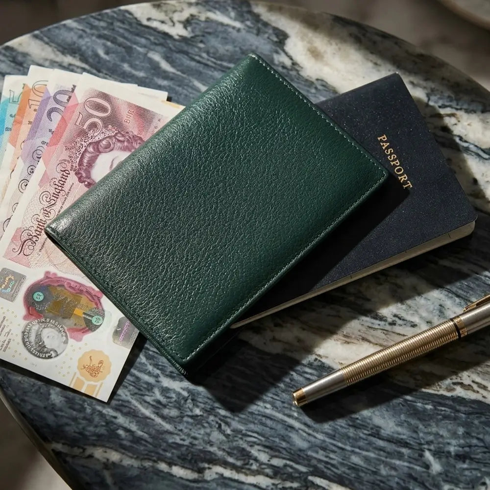 A dark green textured leather passport holder lies on a marble surface, styled with British Pound banknotes, a navy blue passport, and a gold pen.