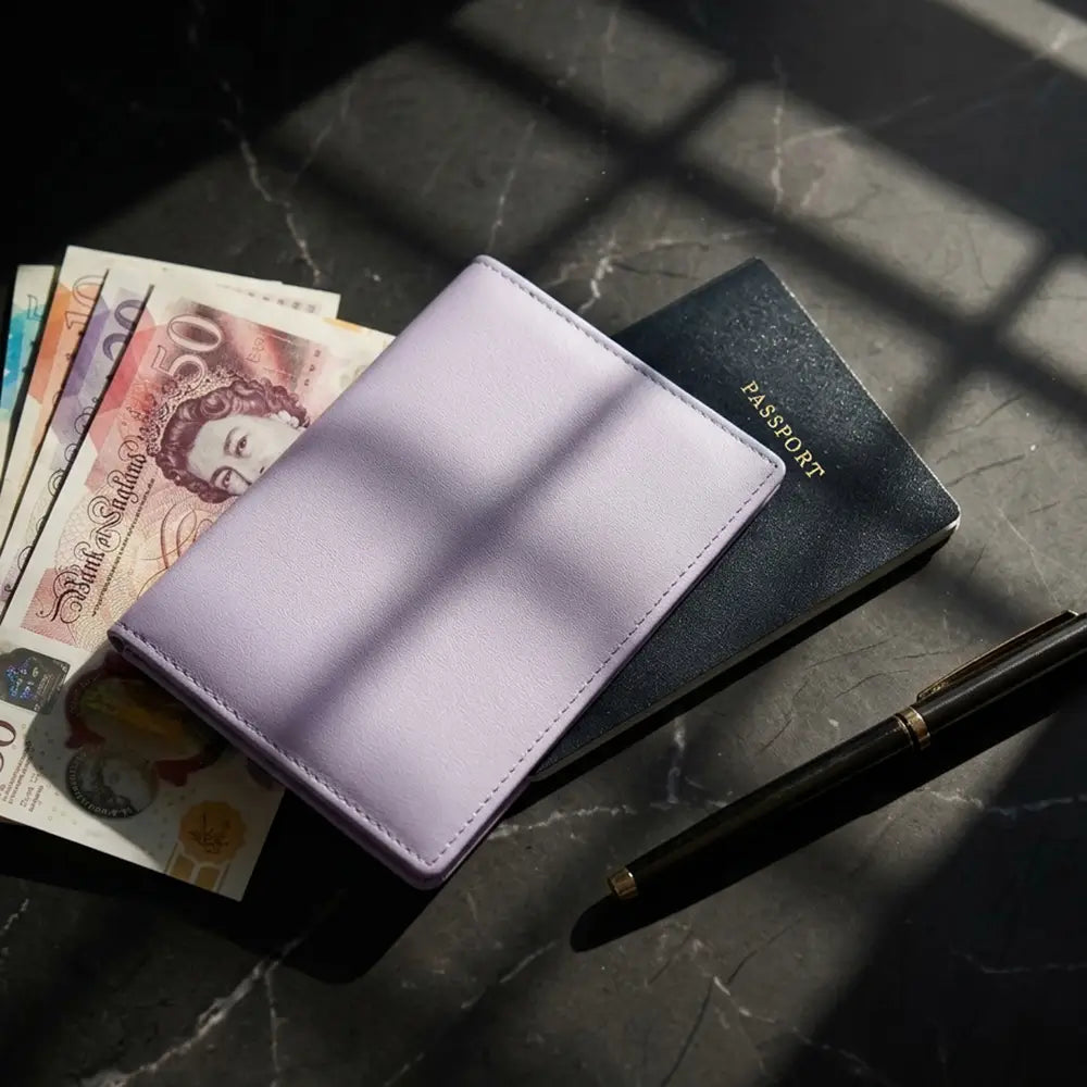 A lilac passport cover lies on top of a dark passport and next to a black pen on a dark marble surface with dramatic window-light shadows.