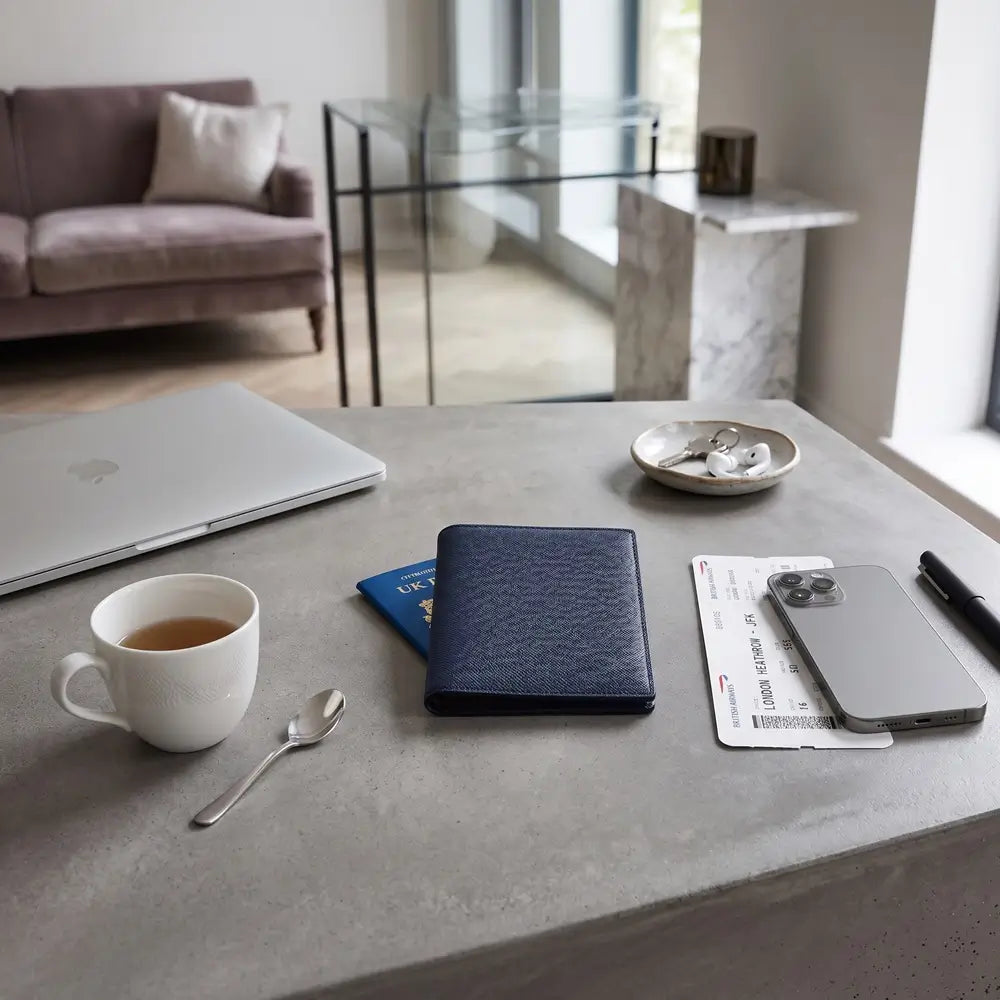 A navy leather passport holder sits on a gray concrete table in a modern living space, surrounded by a white coffee mug, a silver laptop, a smartphone, and other travel documents.
