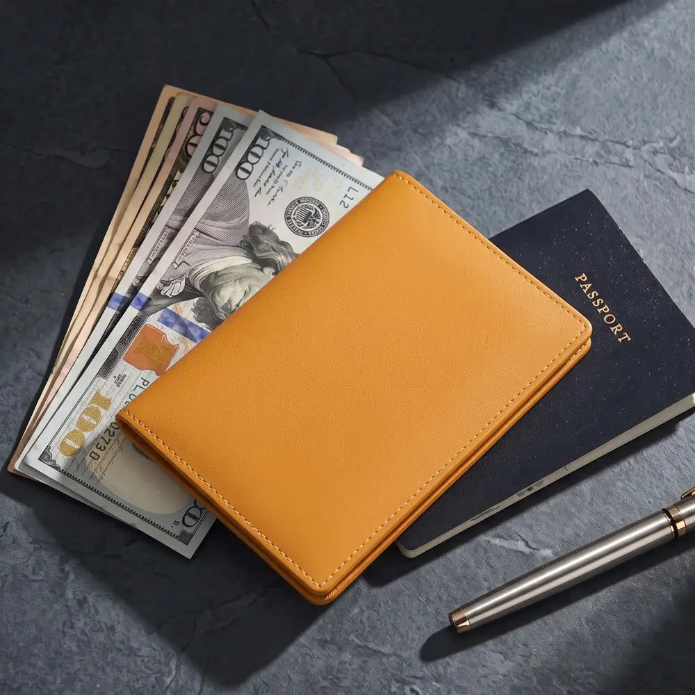 A yellow leather passport holder shown in a styled flat-lay arrangement with a stack of hundred-dollar bills, a black passport, and a silver pen on a dark textured surface.