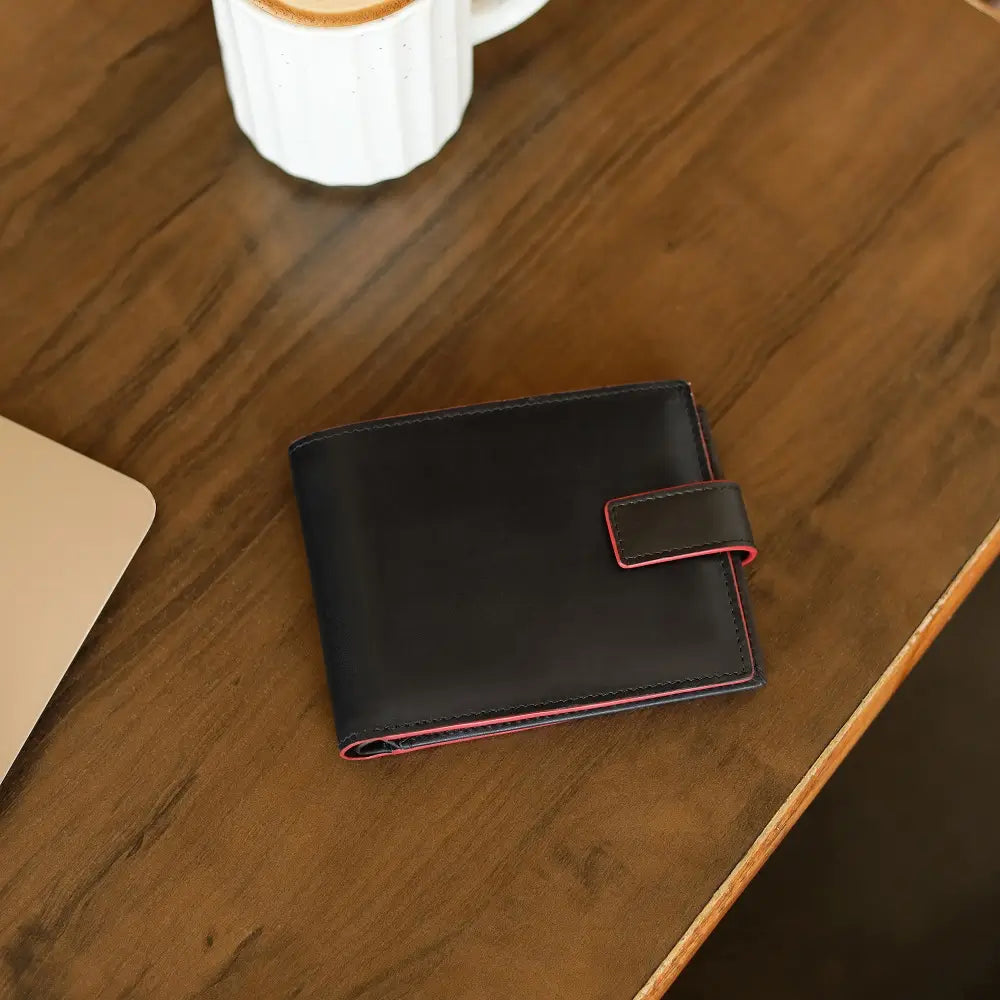 Black leather wallet with red stitching on a wooden surface next to a white mug.