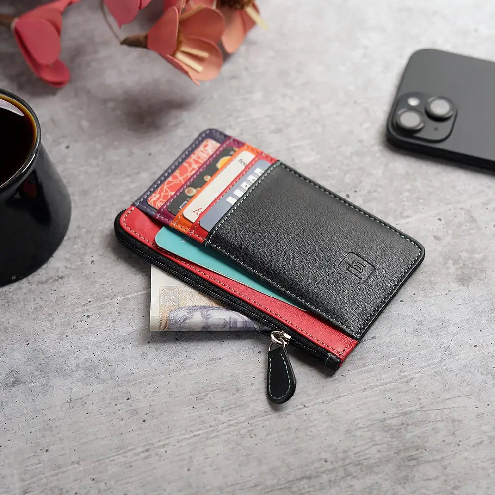 Black card holder purse with red interior holding cards and money on a gray surface with a phone and coffee cup.