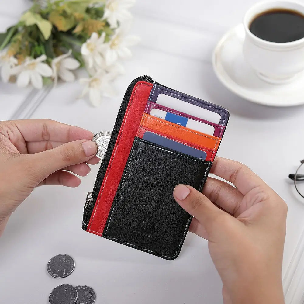 Person holding a black wallet with a red stripe, showing coins and cards on a table with a cup of coffee.