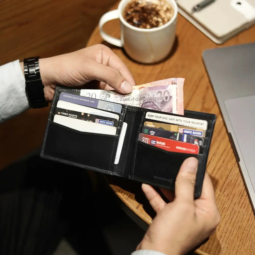 Person holding a black wallet with cards and money on a wooden table with a cup of coffee and laptop.