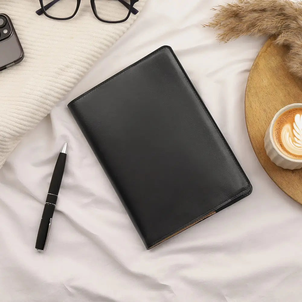 Black leather diary cover on a white surface with a pen, phone, and coffee cup.