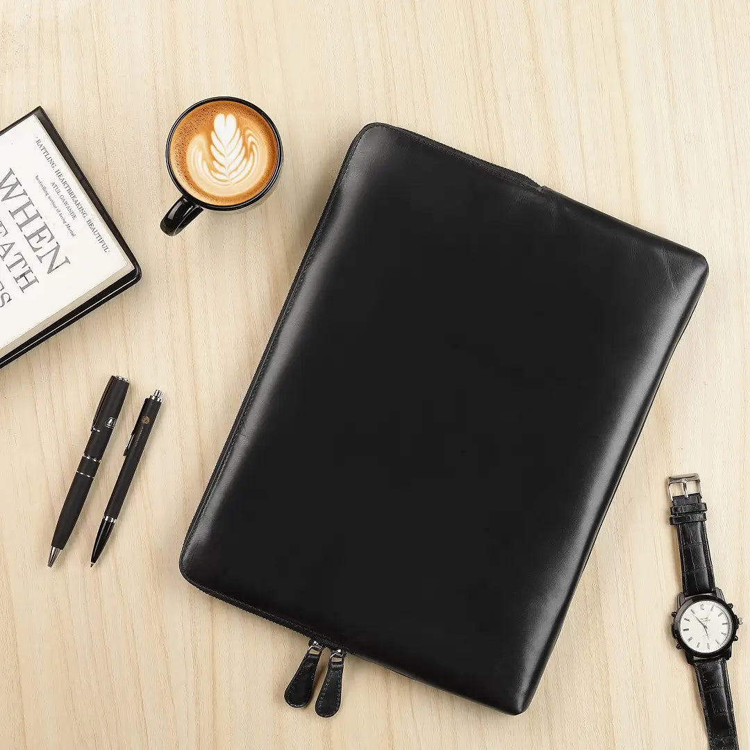 Black leather laptop sleeve on a wooden surface with a coffee cup, pens, and a watch.