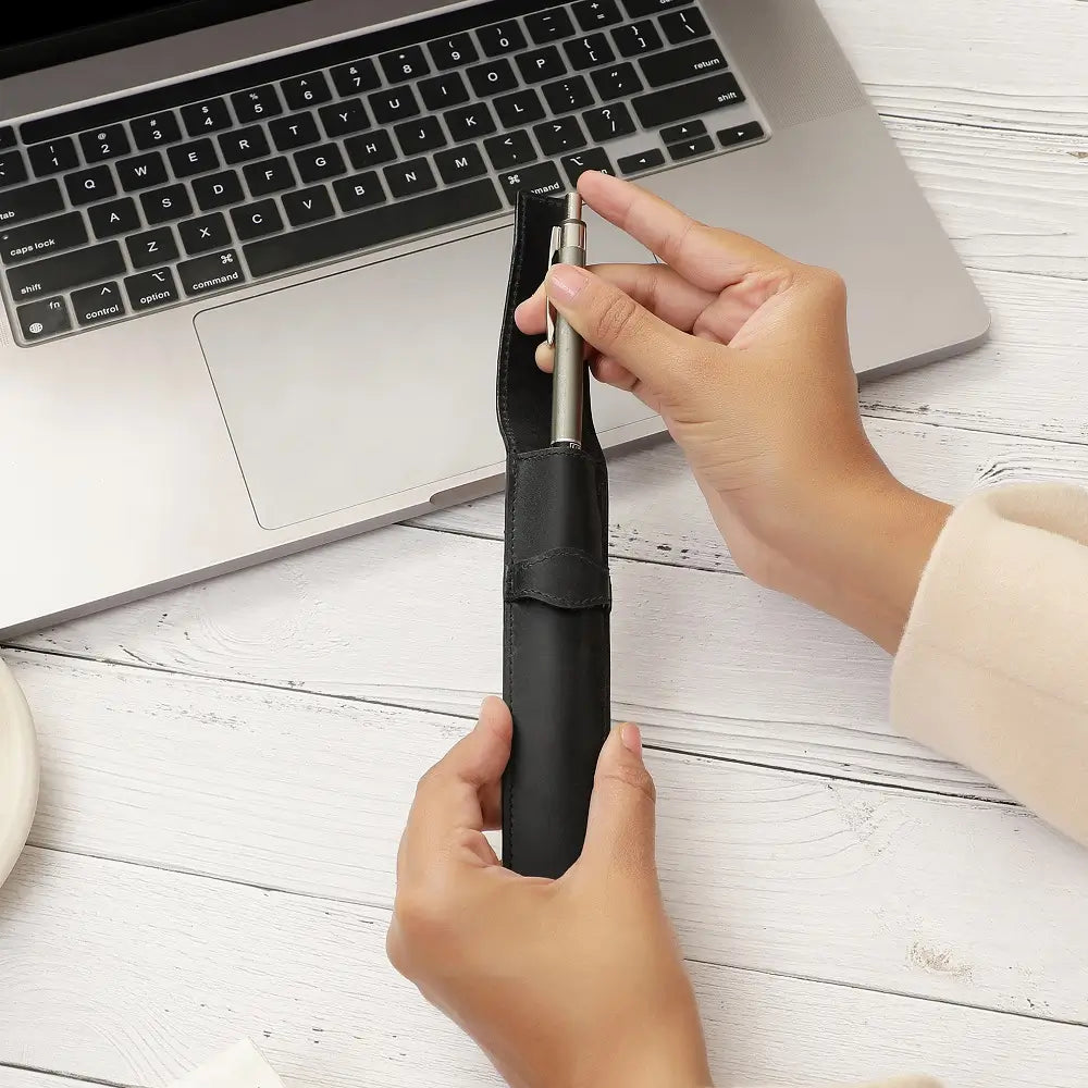 Person using a black leather pen holder on a white surface with a laptop in the background