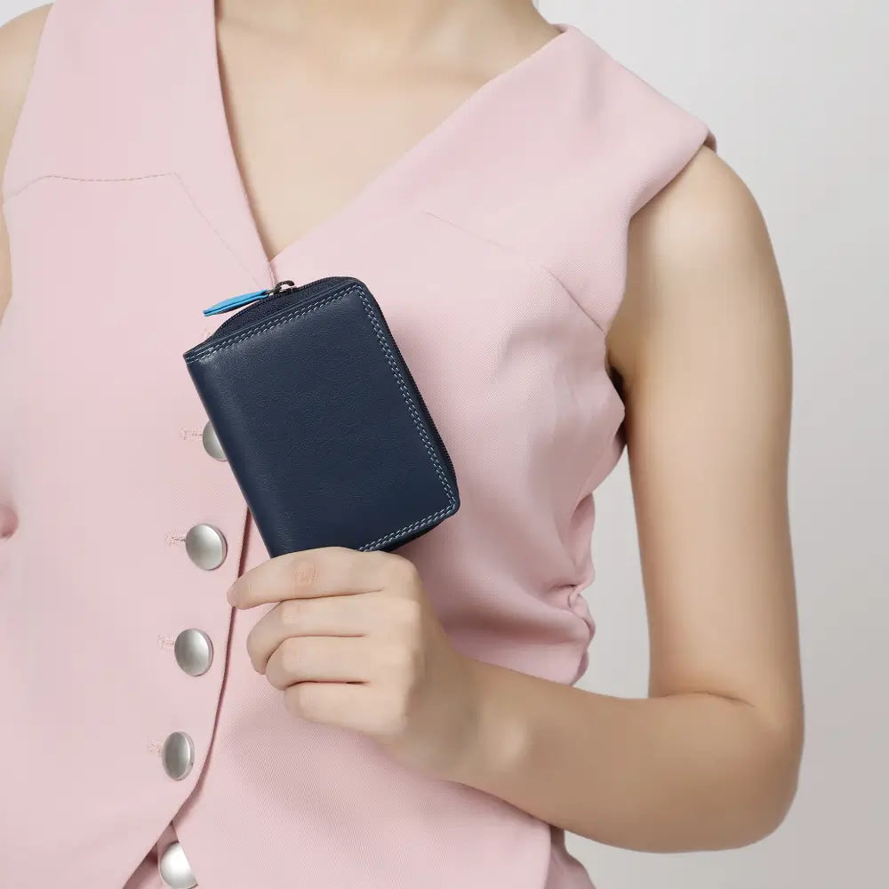 A blue leather zip-around wallet with contrast stitching and a light blue pull tab, styled on a light gray surface with coins, a £20 note, and partial laptop and phone visible.
