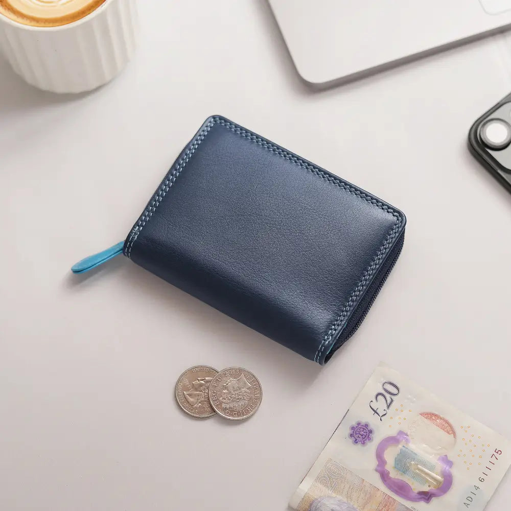 A blue leather zip-around wallet with contrast stitching and a light blue pull tab, styled on a light gray surface with coins, a £20 note, and partial laptop and phone visible.