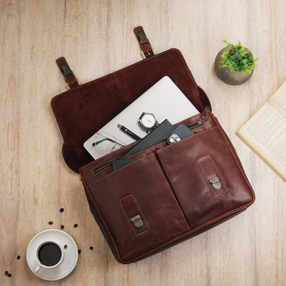 Brown leather laptop bag with items on a wooden surface, next to a cup of coffee and a plant.