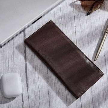Brown leather wallet on a light wooden surface with a laptop and glasses in the background.