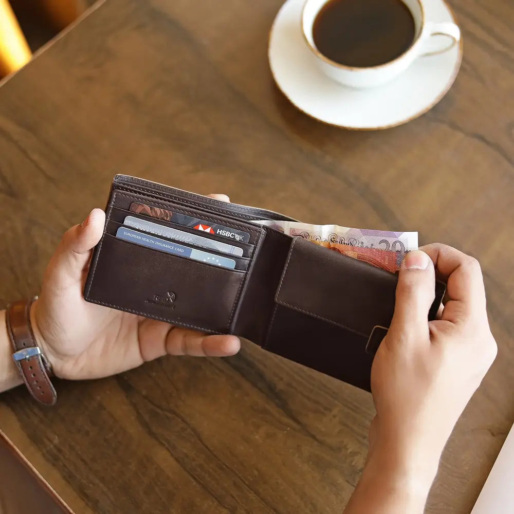 Person holding a brown leather wallet with cards and cash on a wooden table with a cup of coffee.