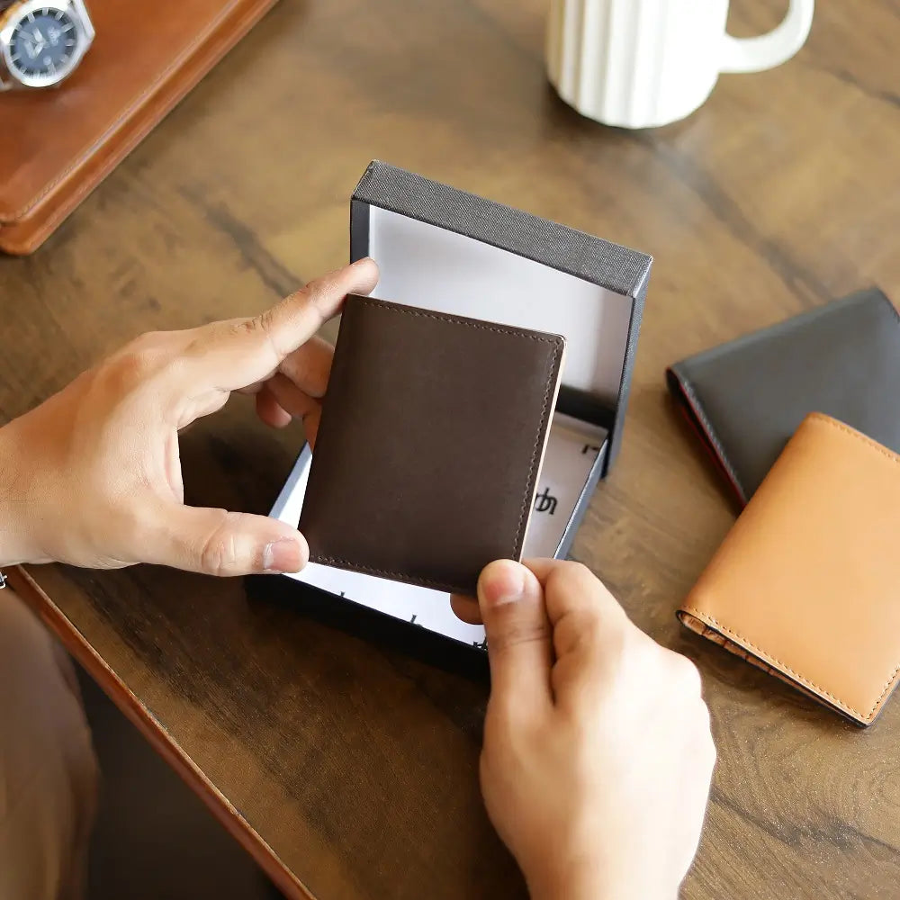 Person holding a brown leather wallet in a box on a wooden table with another wallet and a mug in the background.