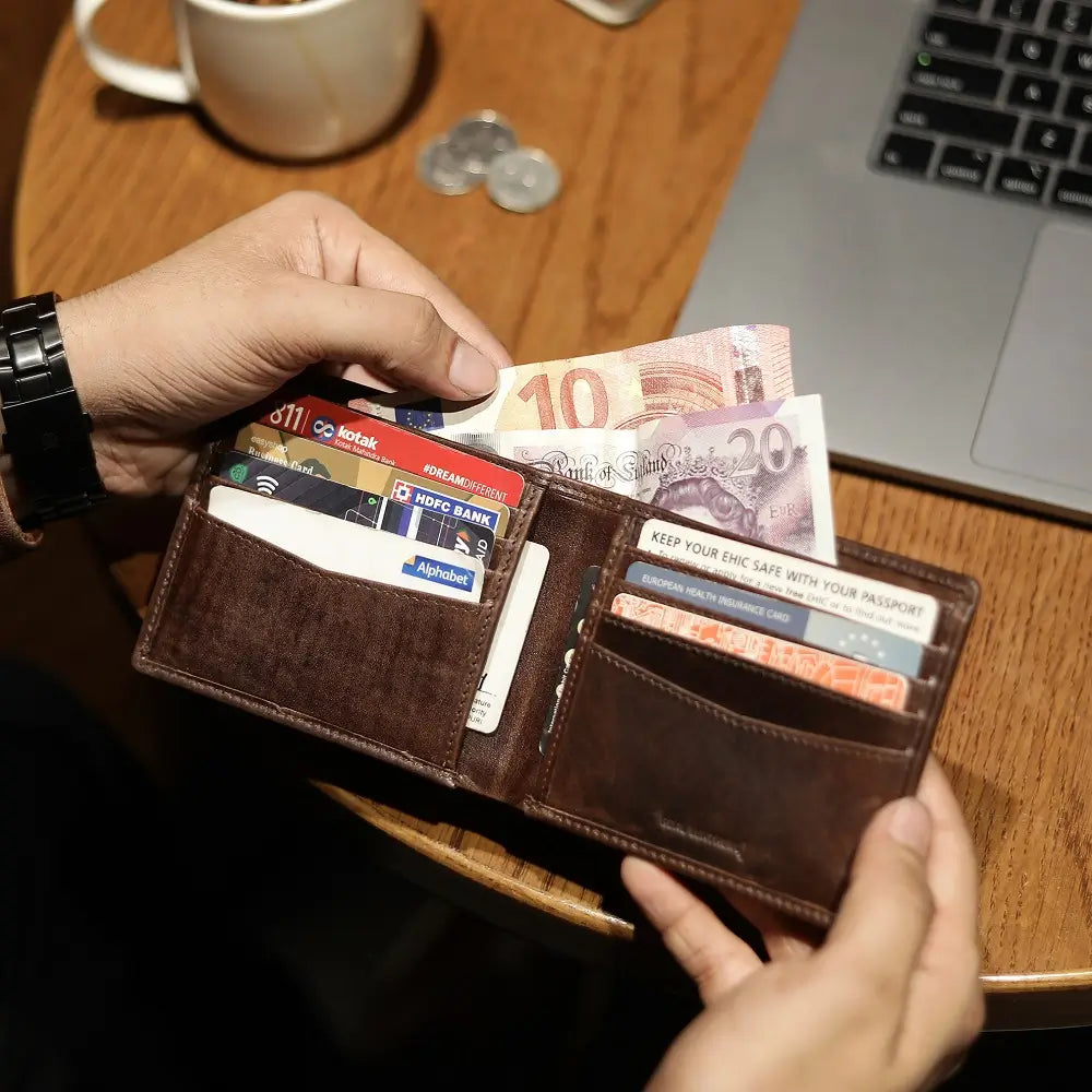 Person holding a brown wallet with currency and cards on a wooden table with a laptop and cup.