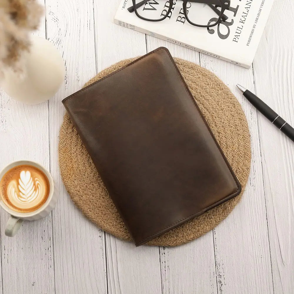 Brown leather diary cover on a textured coaster with a cup of coffee, pen, and magazine in the background.