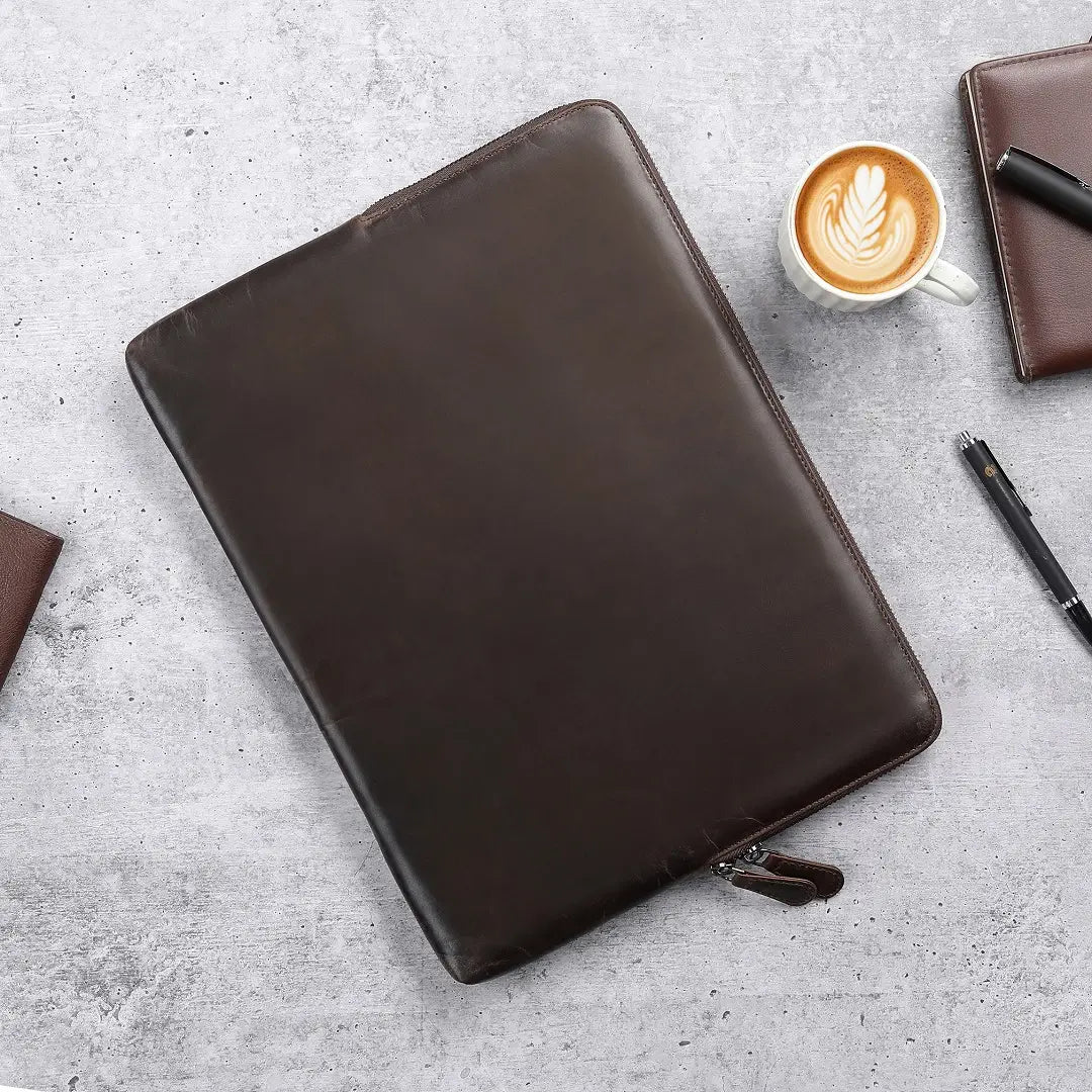 Brown leather laptop sleeve on a gray surface with a cup of coffee and pen.