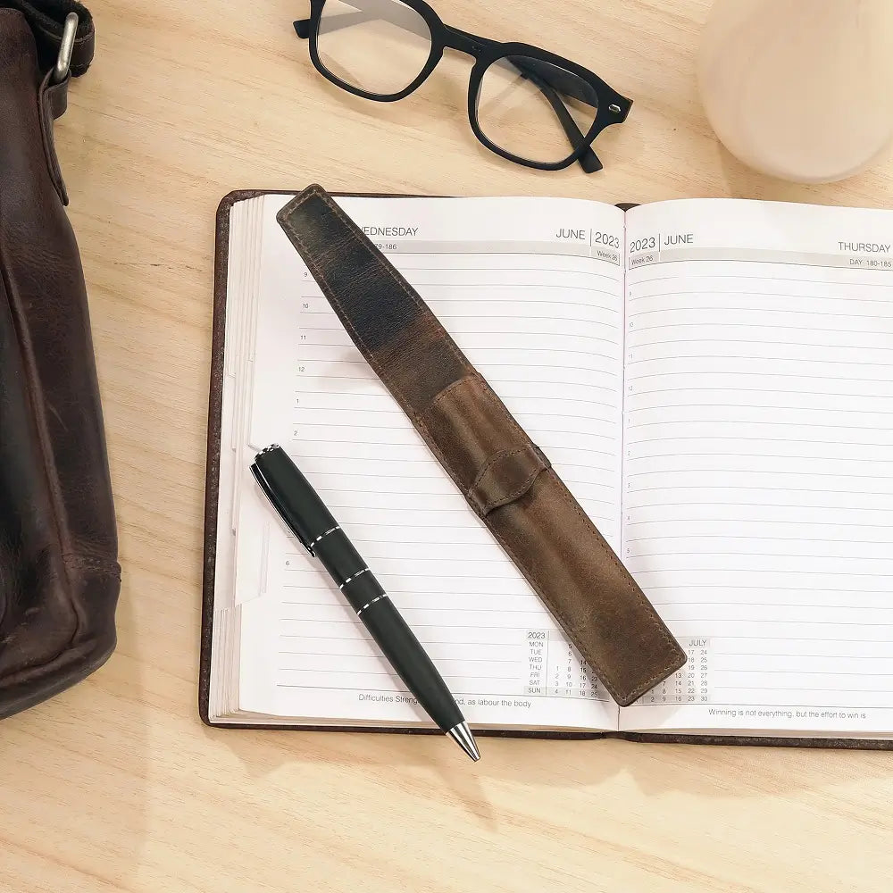 Open planner with leather pen holder and black pen on a wooden surface, with glasses and brown bag in the background.