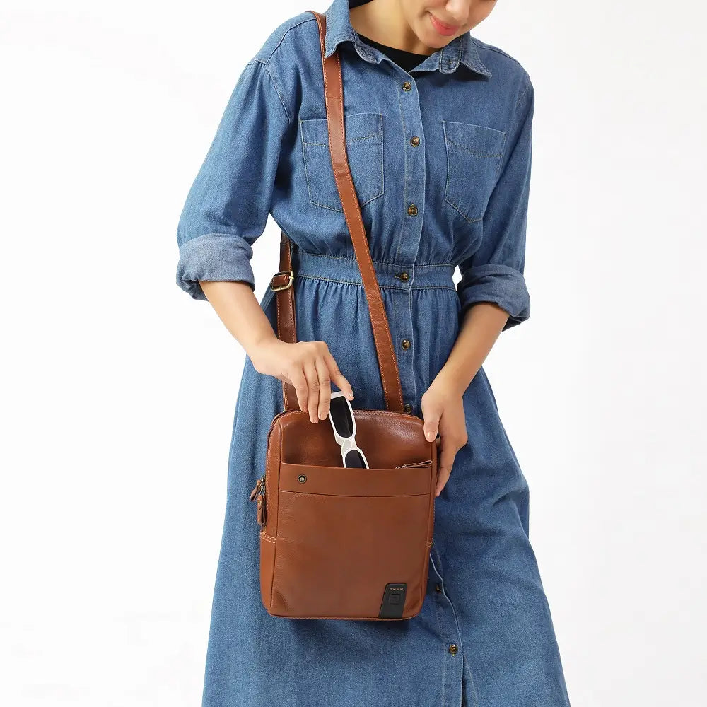 Person wearing a denim dress holding a tan messenger bag on a white background