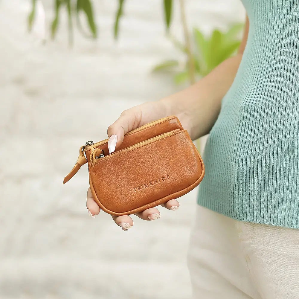 Person holding a brown leather pouch with 'PRIMEHIDE' branding against a blurred natural background