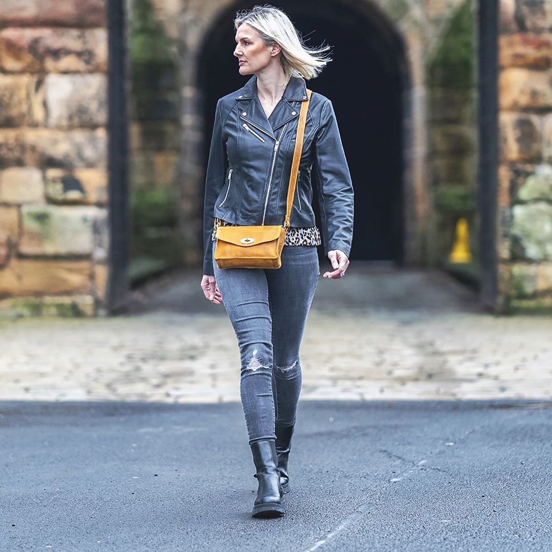 a woman in a black leather jacket with a yellow leather handbag. she is leaning against a stone wall