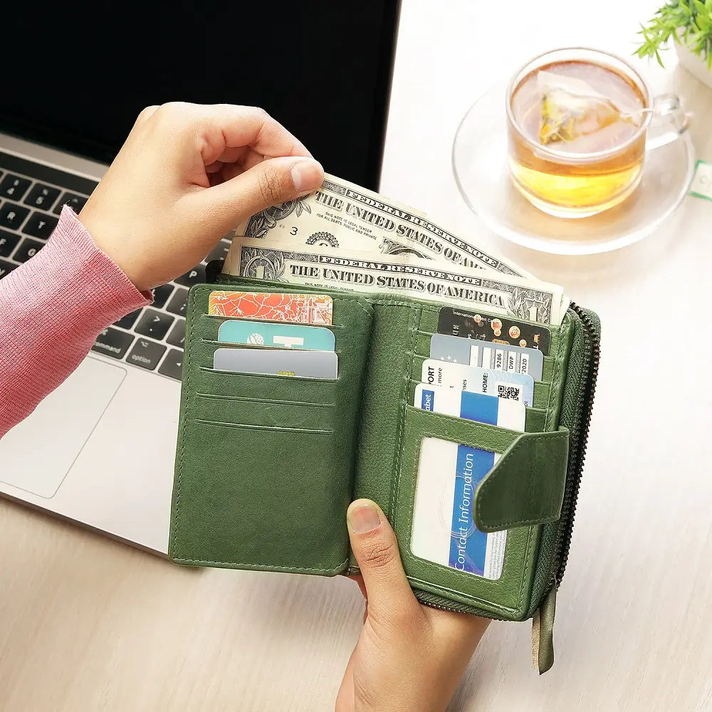 Green wallet with money and cards held by a hand on a desk with a laptop and tea.