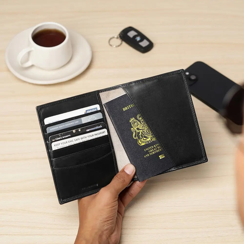 Black leather wallet with multiple card slots and a passport holder, held by a hand on a wooden surface with a cup of tea and phone in the background.