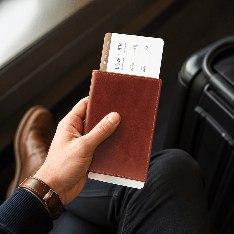 Hand holding a brown leather passport holder with a passport and boarding pass inside, sitting next to a suitcase.