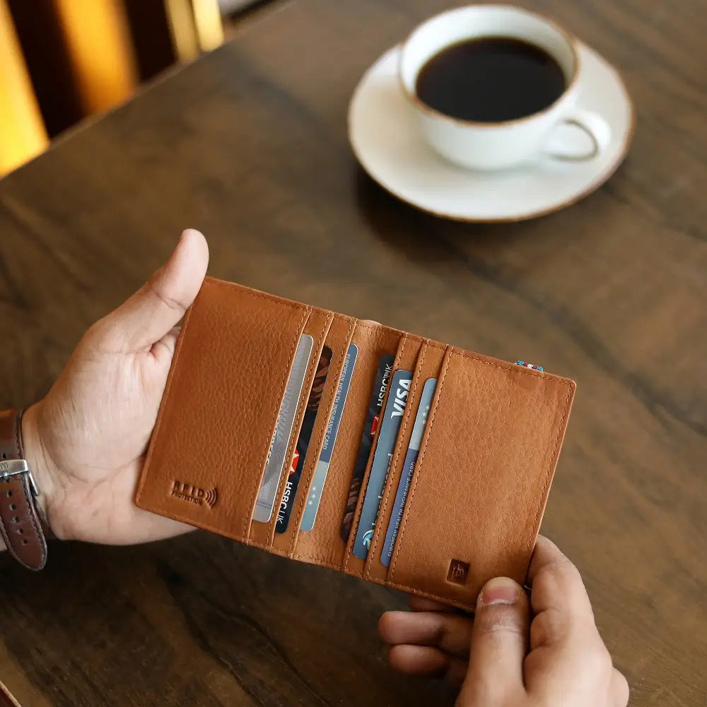 Cognac leather wallet with cards held by a hand on a wooden table with a cup of coffee.