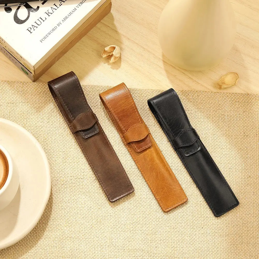 Three leather pen holders in brown, tan, and black on a textured surface with a book and cup in the background.
