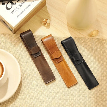 Three leather pen holders in brown, tan, and black on a textured surface with a book and cup in the background.