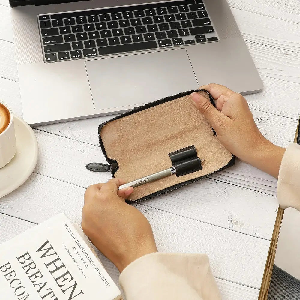 Person holding a pen case with a pen on a desk next to a laptop and book.