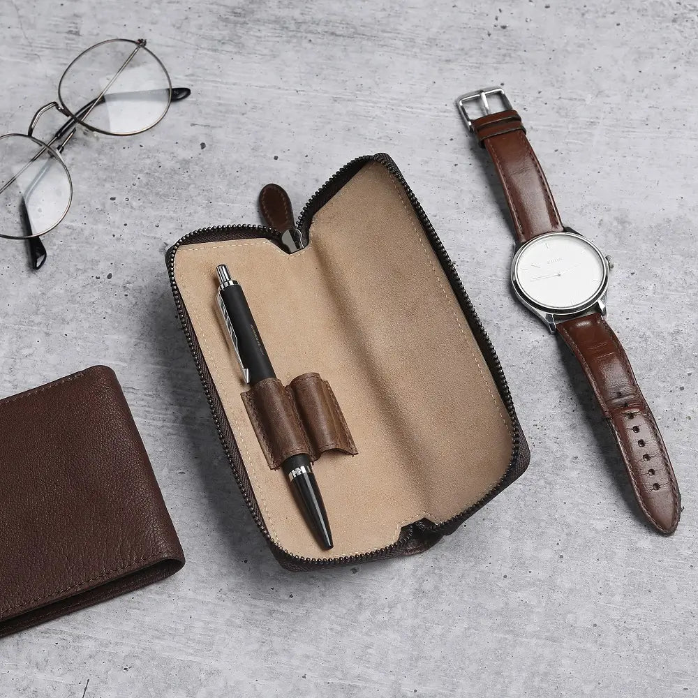 Brown leather wallet, pen case with pen, glasses, and watch on a gray surface