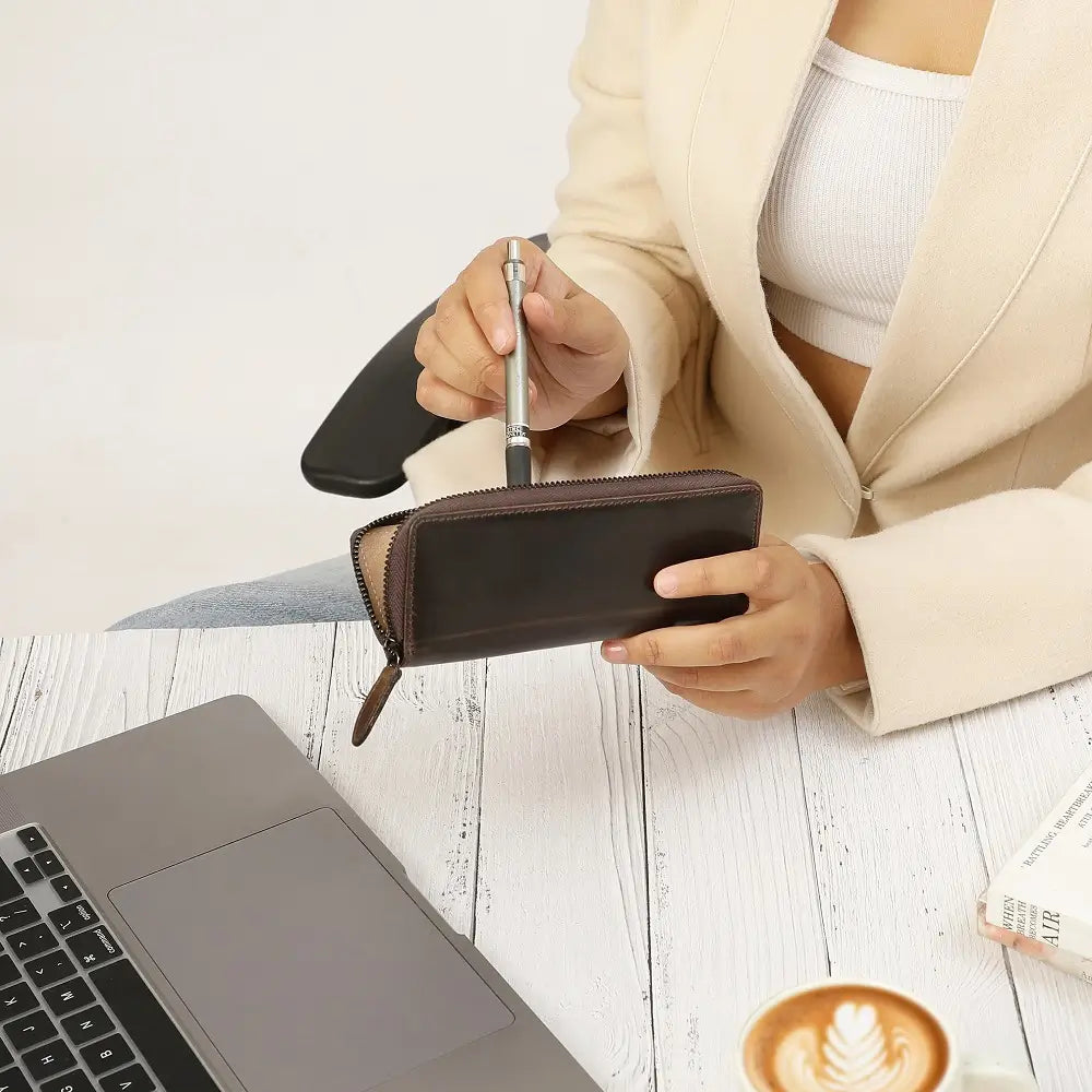Person holding a pen and pen case next to a laptop on a wooden surface