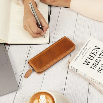 Tan leather pen case on a wooden surface with a notebook, pen, and coffee.