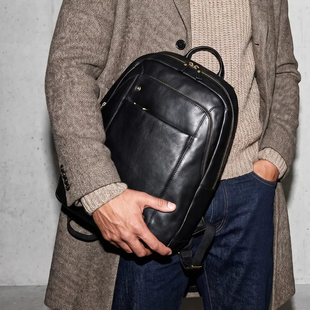 A person wearing a brown tweed blazer and blue jeans holds a black leather backpack against their side.