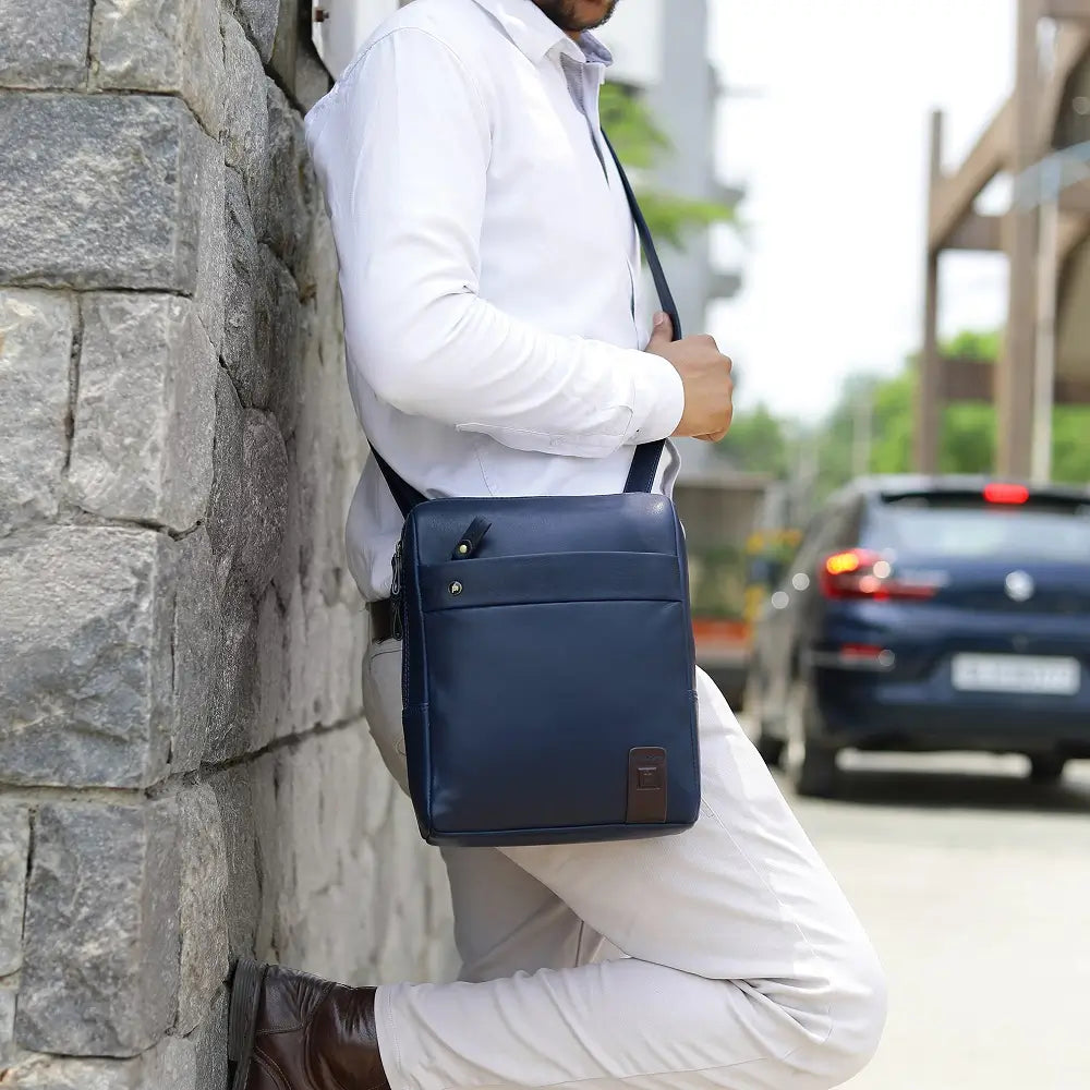 Man leaning against a stone wall wearing a white shirt and dark pants with a navy messenger  bag.
