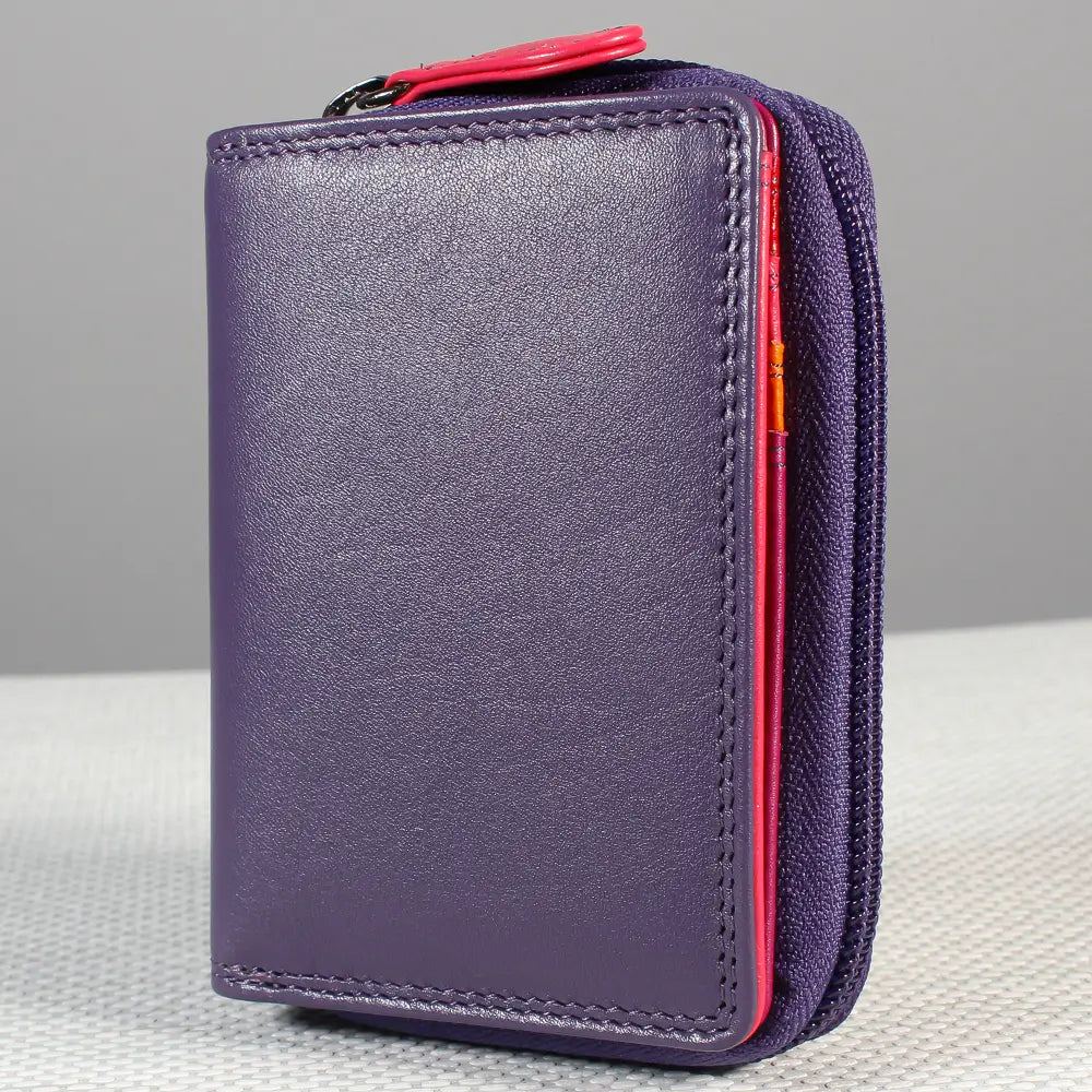 A purple leather zip-around wallet with a bright pink zipper pull and pink inner lining, shown standing upright on a light textured surface against a gray background.