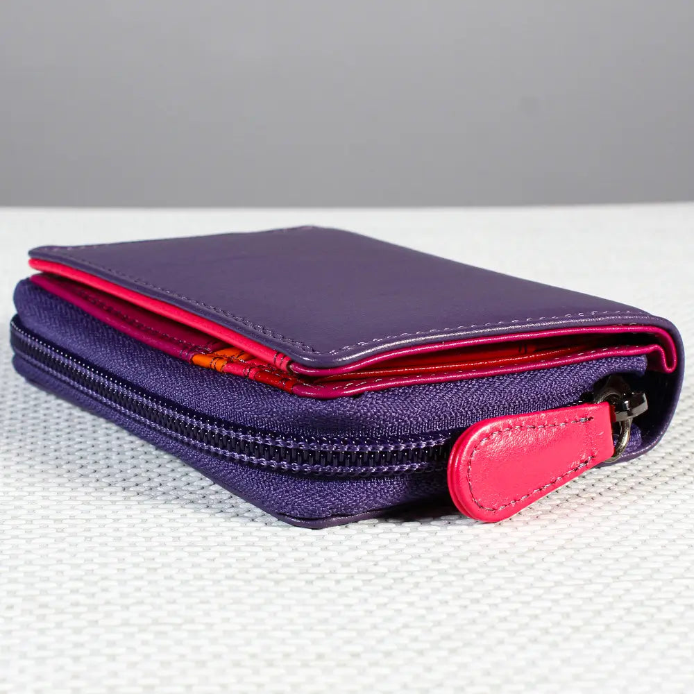 A purple leather zip-around wallet with a bright pink interior lining and matching pink leather zipper pull, shown on a textured white surface against a gray background.