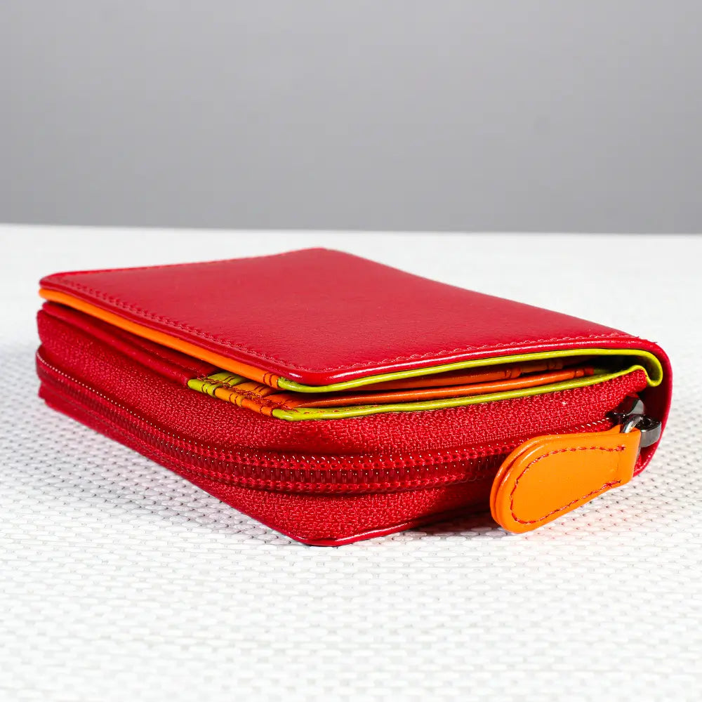 A red zippered wallet with card slots, featuring a bright orange leather pull tab and a thin yellow-green accent strip along the edge, set on a white textured surface against a gray background.