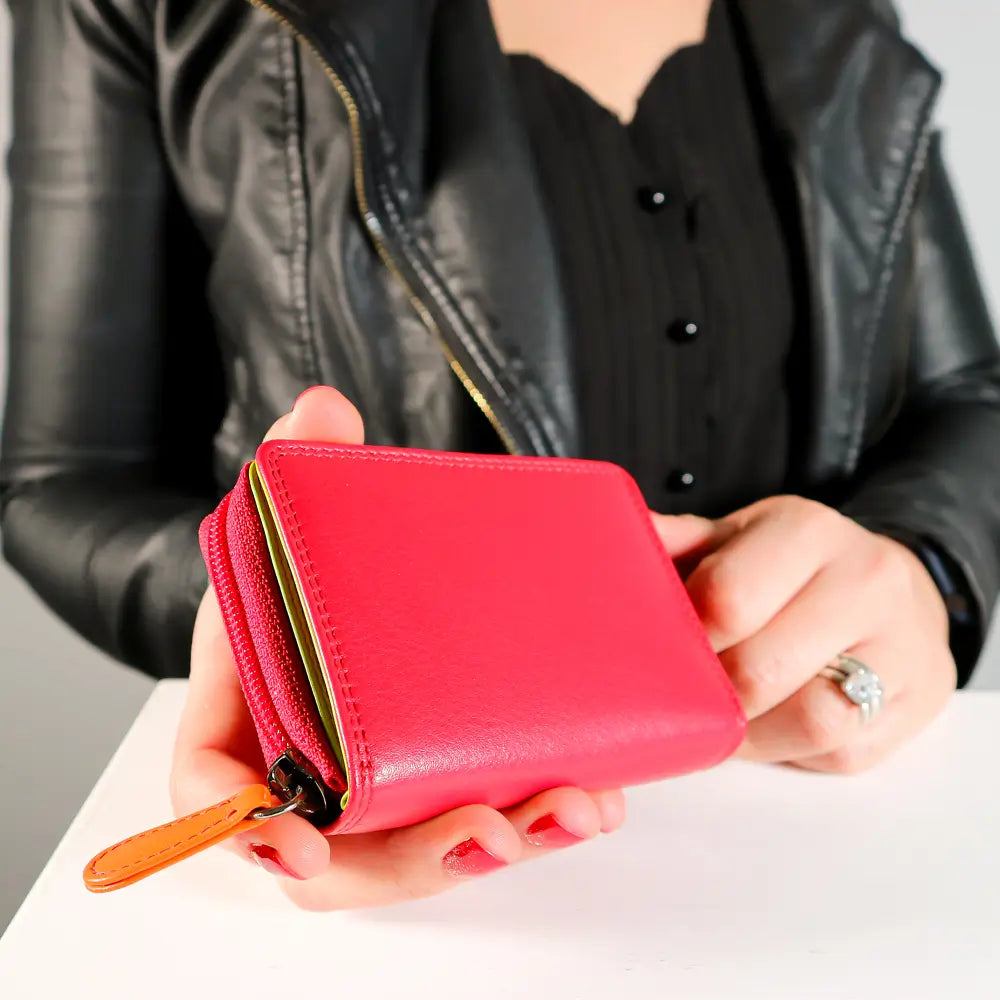 A person in a black leather jacket holds a small bright pink zip-around wallet with an orange leather pull tab and gold-tone zipper detail.