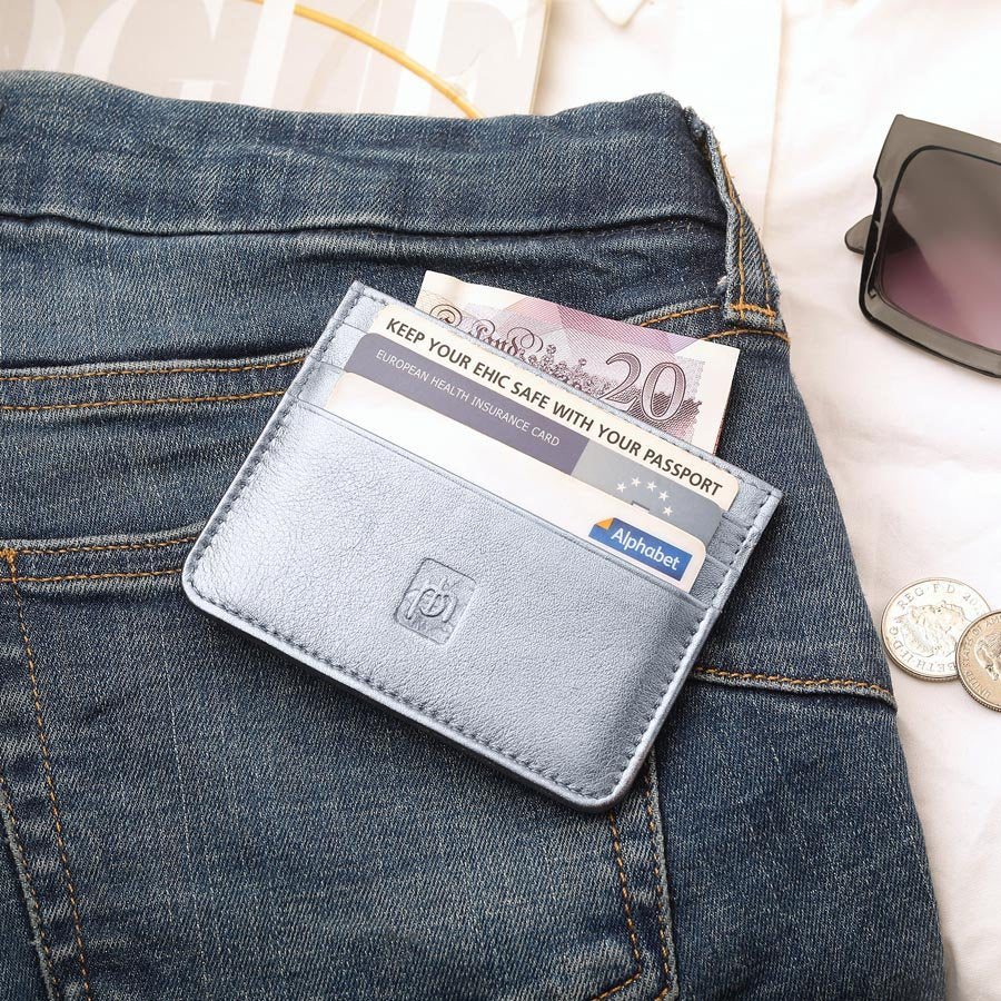 Silver personalised leather card holder filled with cash and cards, styled on a pair of blue jeans alongside sunglasses and coins.