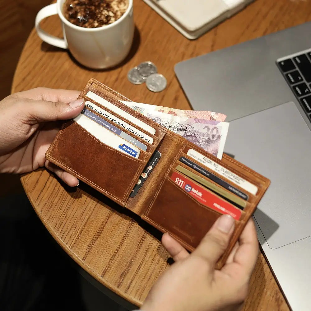Tan leather wallet with cards and money held by hands on a wooden table with a laptop and coffee.